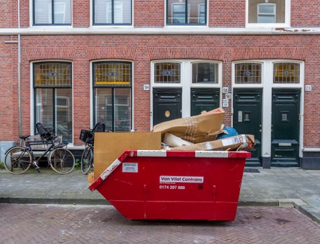 A large red rubbish skip positioned on a residential pavement in front of a row of brick townhouses with white-framed windows and dark green front doors. The skip contains various waste materials, including a large rolled-up cardboard box, a crumpled brown paper bag, and other mixed debris, indicating clearance or disposal activity typical of a rubbish removal service. To the left of the skip, two bicycles are parked close to the buildings, with one bike secured to a metal railing. The brick facades and pavement suggest an urban residential area, possibly within Cranford or nearby. The scene is well-lit with natural daylight, emphasizing the textures of the waste, brickwork, and bicycles, and reflecting the typical environment for garden or rubbish clearance services offered by Garden Clearance Cranford, often involving waste collection from urban residential properties.