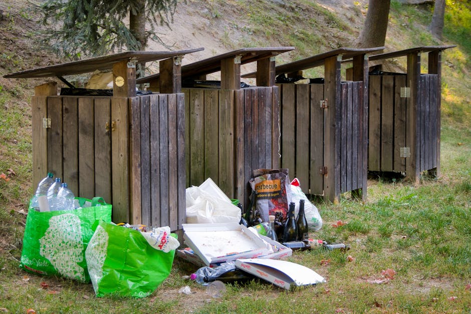 An aerial view of a large outdoor waste accumulation area filled with a dense layer of mixed rubbish, including plastic bottles, paper, cardboard, and other household waste materials. The waste appear