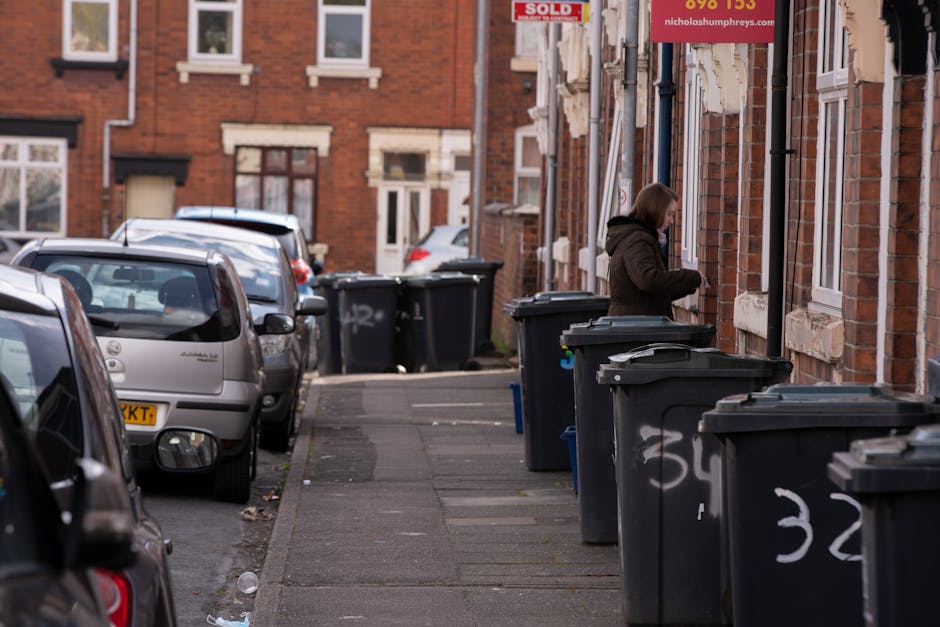 A large collection of mixed household waste and recycling including paper, cardboard boxes, plastic bags, black rubbish sacks, and loose packaging, situated on a paved area near a parking space. The w